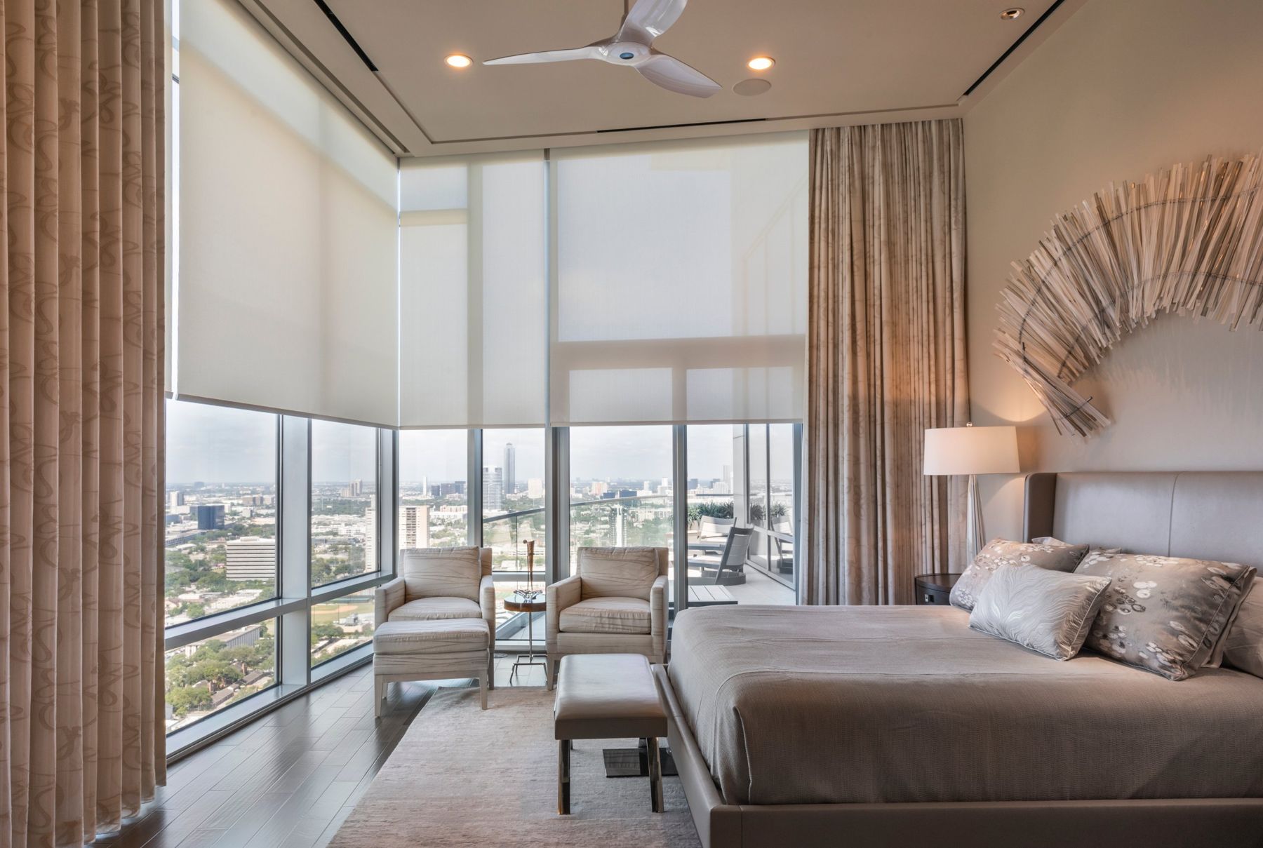 Corner primary bedroom with sheer motorized roller shades over floor-to-ceiling glass and a downtown view