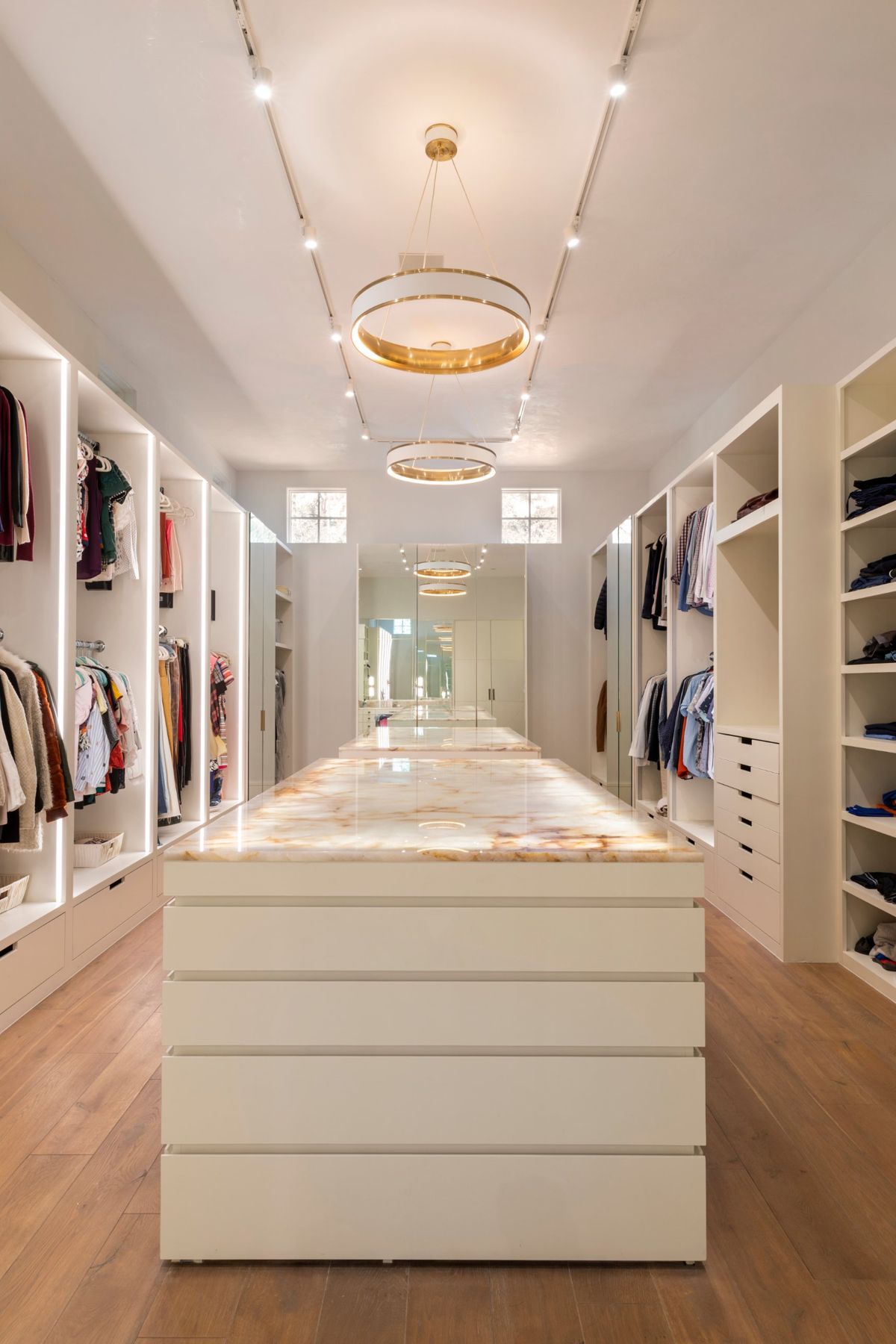 Primary closet with lit hanging bays, brass ring pendants and a backlit onyx island