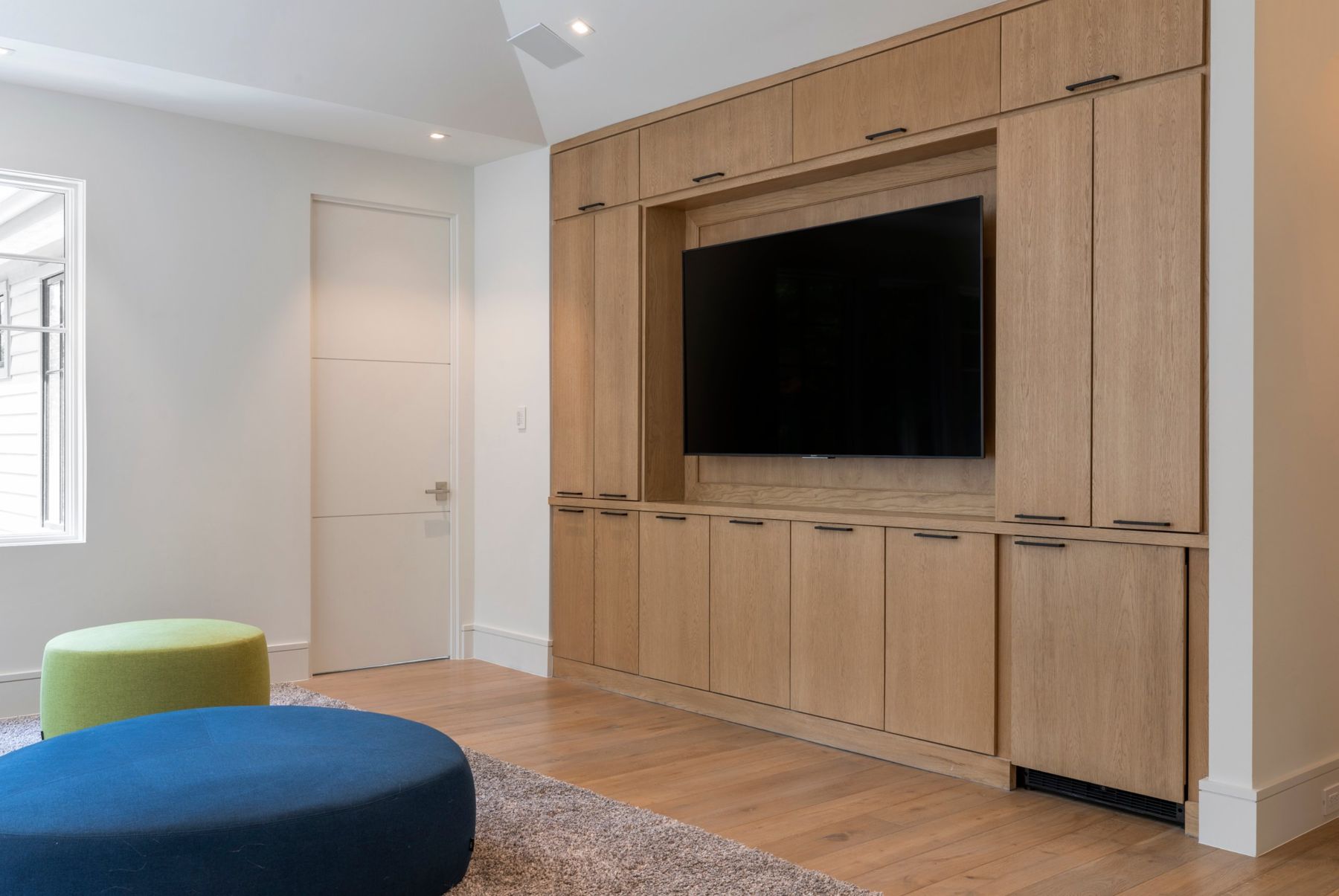 Game room with a TV set into white oak cabinetry and colorful ottomans
