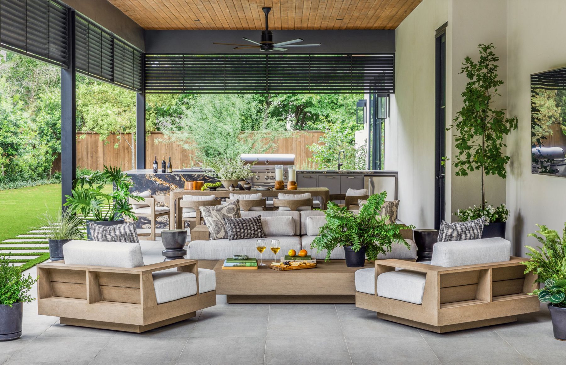 Covered outdoor living room with an outdoor TV, ceiling fan and summer kitchen opening onto the lawn