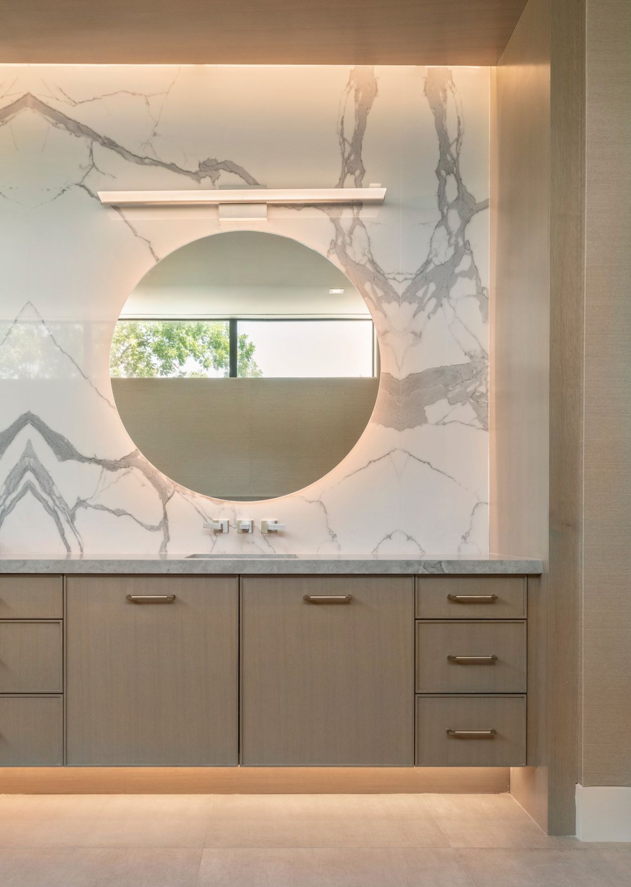 Primary bath vanity with a backlit round mirror on a book-matched stone wall